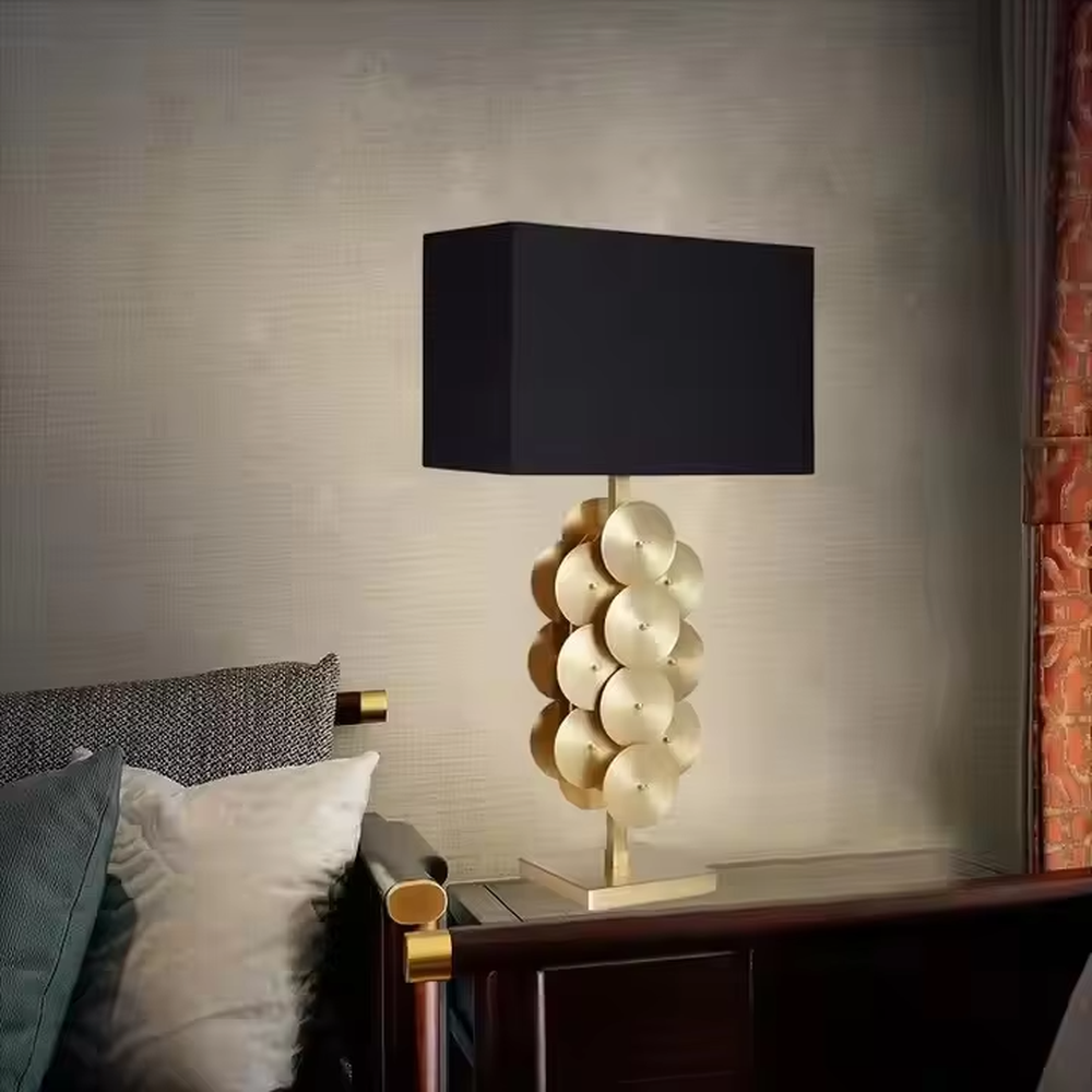 This is a luxury-style black and gold table lamp, with a golden disc-stacked base and a black square fabric lampshade, featuring a minimalist and grand design with strong luxury texture. It is used for local desktop lighting and decoration, creating a luxury and modern atmosphere. It is suitable for luxury-style home bedroom bedside tables, high-end hotel guest rooms, modern minimalist living room side tables and other places.