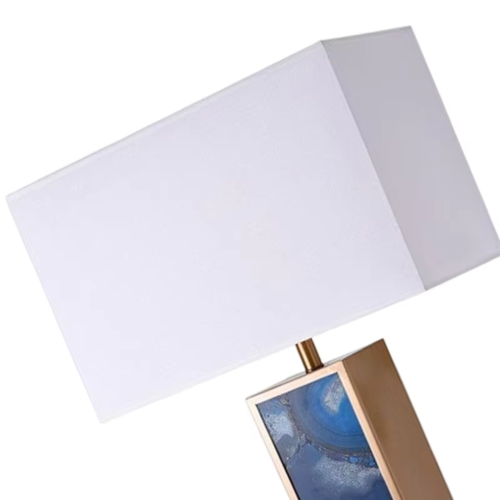 This is a luxury artistic agate-textured table lamp, with a golden metal frame inlaid with blue agate texture decoration and a white square fabric lampshade, featuring a minimalist and grand design with both artistic sense and luxury texture. It is used for local desktop lighting and decoration, creating a luxury artistic atmosphere. It is suitable for luxury-style home bedroom bedside tables, high-end hotel guest rooms, art exhibition hall display tables and other places.