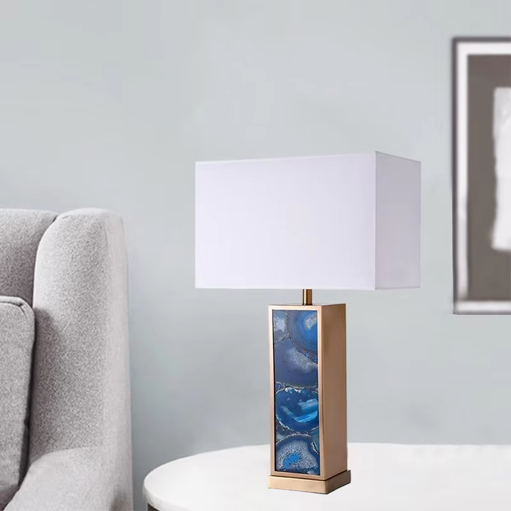This is a luxury artistic agate-textured table lamp, with a golden metal frame inlaid with blue agate texture decoration and a white square fabric lampshade, featuring a minimalist and grand design with both artistic sense and luxury texture. It is used for local desktop lighting and decoration, creating a luxury artistic atmosphere. It is suitable for luxury-style home bedroom bedside tables, high-end hotel guest rooms, art exhibition hall display tables and other places.
