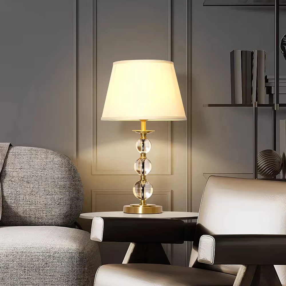 This is a light luxury exquisite crystal ball column table lamp, with a golden metal base and connectors, paired with transparent crystal ball columns and a beige fabric lampshade. It features a light luxury and exquisite design, with warm and soft lighting. It is used for local desktop lighting and decoration, creating a light luxury elegant atmosphere. Suitable for light luxury-style bedroom bedside tables, modern minimalist living room side tables, high-end hotel guest rooms and other places.