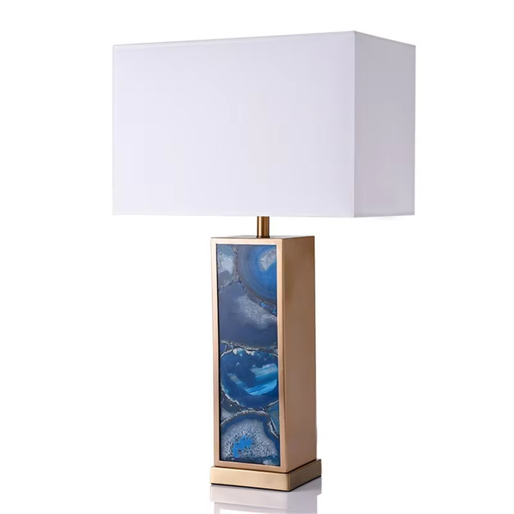 This is a luxury artistic agate-textured table lamp, with a golden metal frame inlaid with blue agate texture decoration and a white square fabric lampshade, featuring a minimalist and grand design with both artistic sense and luxury texture. It is used for local desktop lighting and decoration, creating a luxury artistic atmosphere. It is suitable for luxury-style home bedroom bedside tables, high-end hotel guest rooms, art exhibition hall display tables and other places.