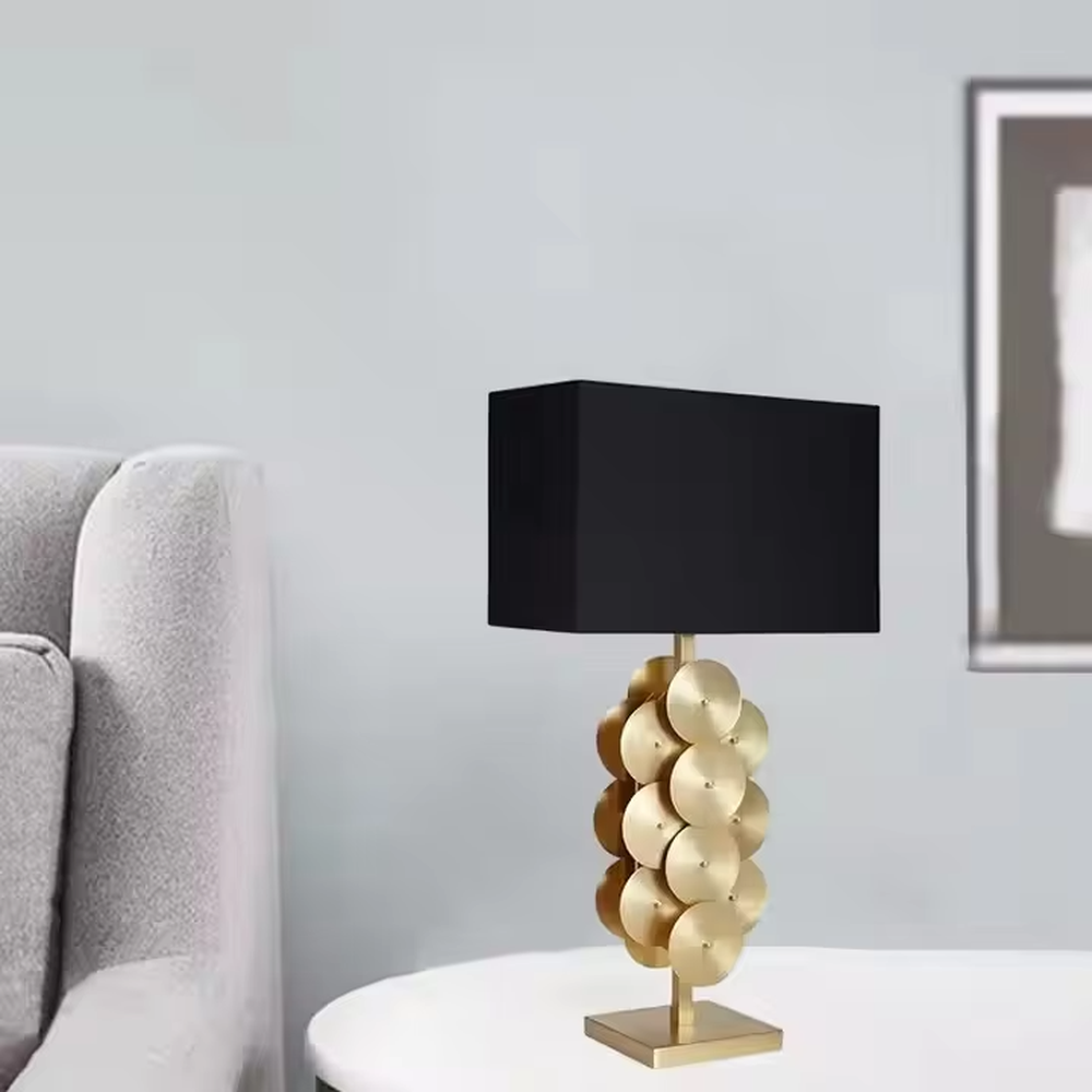 This is a luxury-style black and gold table lamp, with a golden disc-stacked base and a black square fabric lampshade, featuring a minimalist and grand design with strong luxury texture. It is used for local desktop lighting and decoration, creating a luxury and modern atmosphere. It is suitable for luxury-style home bedroom bedside tables, high-end hotel guest rooms, modern minimalist living room side tables and other places.