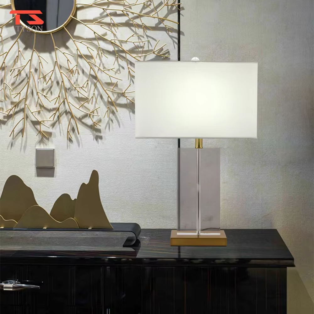This modern light luxury table lamp features an off-white fabric lampshade (trimmed with a thin gold edge), paired with a transparent crystal square column lamp pole and a gold metal base. The overall style is simple and elegant, with soft eye-protective light, serving both lighting and decorative purposes, suitable for modern light luxury style spaces such as bedrooms, living rooms, and hotel guest rooms.