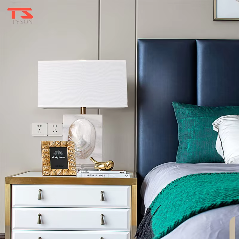 This light luxury modern table lamp features a light-colored stone base with shell relief, paired with gold metal connectors and an off-white fabric rectangular lampshade. It serves both lighting and decorative purposes, ideal for light luxury spaces such as bedroom bedside tables, living room side tables, and hotel guest rooms.