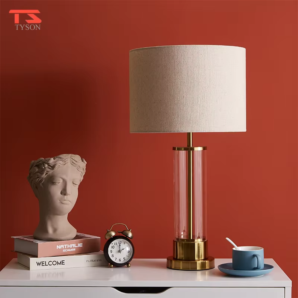 This modern light luxury table lamp features a transparent glass lamp body, paired with gold metal connectors and base, and an off-white fabric cylindrical lampshade that transmits light softly. It serves both lighting and decorative purposes, ideal for light luxury spaces such as bedroom bedside tables, living room side tables, and hotel guest rooms.