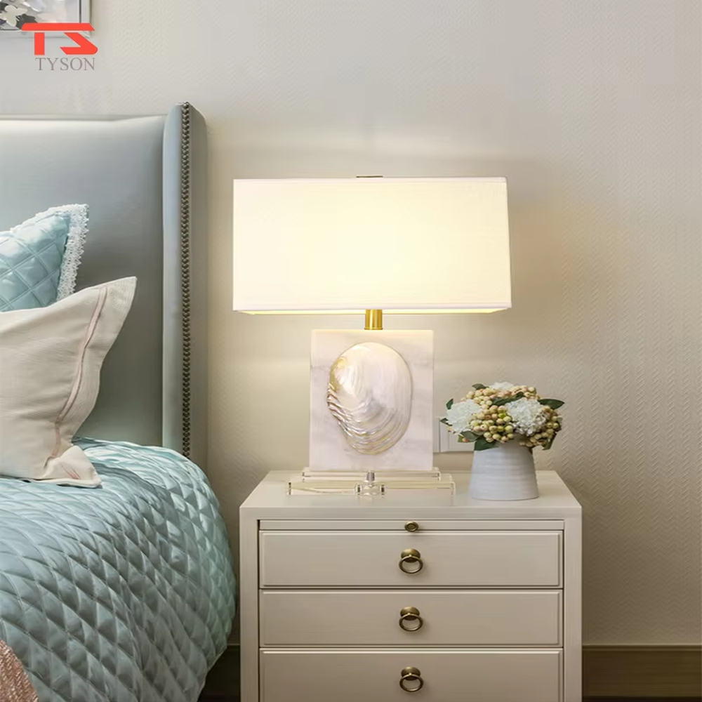 This light luxury modern table lamp features a light-colored stone base with shell relief, paired with gold metal connectors and an off-white fabric rectangular lampshade. It serves both lighting and decorative purposes, ideal for light luxury spaces such as bedroom bedside tables, living room side tables, and hotel guest rooms.