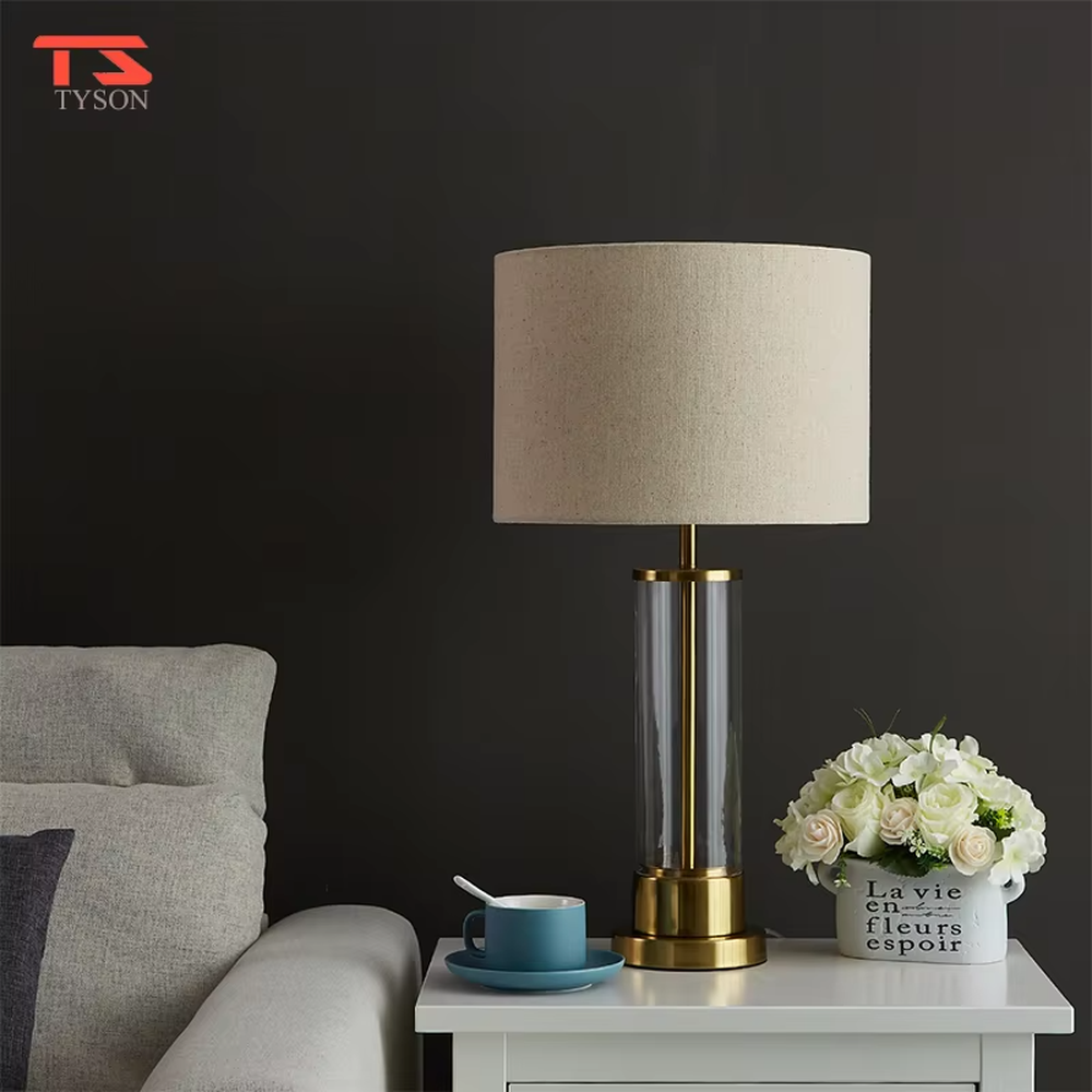 This modern light luxury table lamp features a transparent glass lamp body, paired with gold metal connectors and base, and an off-white fabric cylindrical lampshade that transmits light softly. It serves both lighting and decorative purposes, ideal for light luxury spaces such as bedroom bedside tables, living room side tables, and hotel guest rooms.