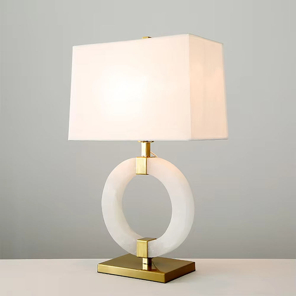 Modern light luxury style table lamp with a white natural stone/acrylic ring paired with gold metal brackets and base, and a rectangular white fabric lampshade. It emits soft and even light, combining decorativeness and practicality, suitable for lighting and decoration in spaces such as bedroom nightstands, living room side tables, and hotel guest rooms.