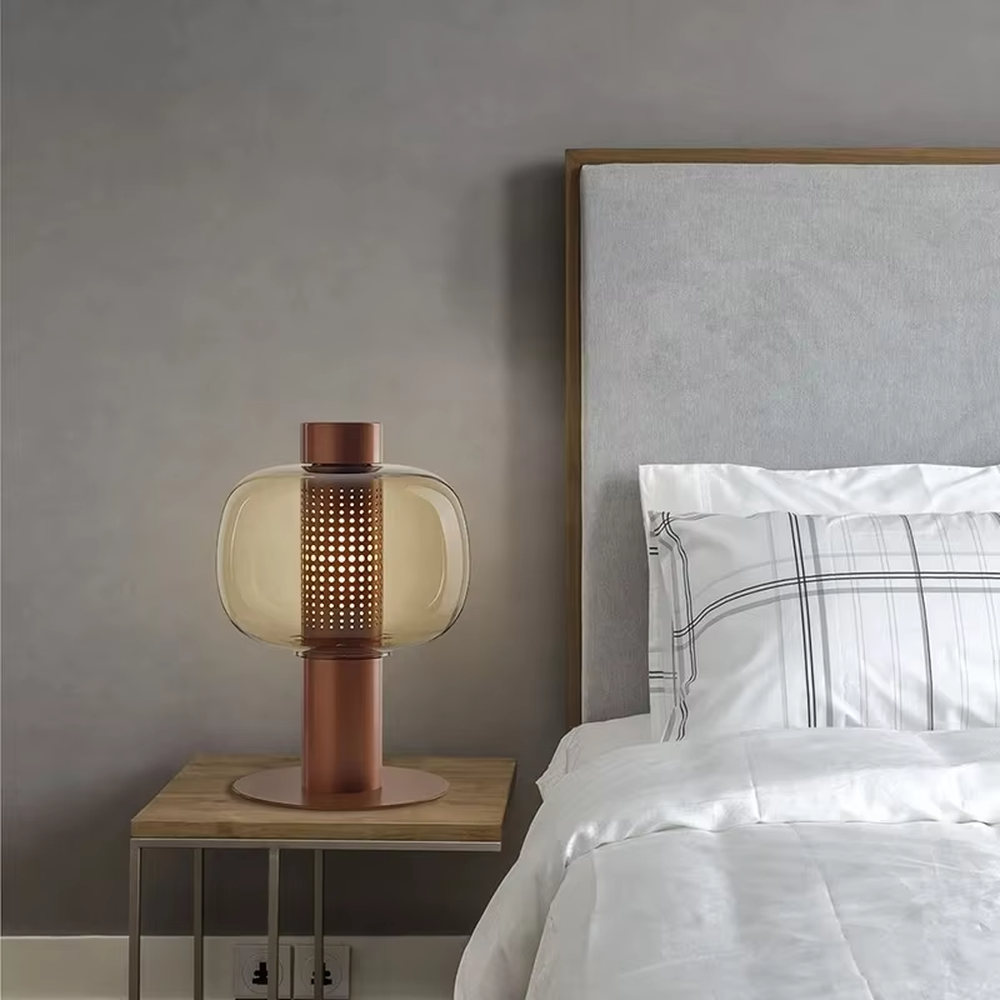 This modern minimalist table lamp features a copper-colored metal frame, paired with an oval smoked glass lampshade and a perforated metal lamp core. Its shape combines roundness with industrial details, and light passes through the perforations and glass to create soft, dappled light and shadow. It serves both lighting and decorative purposes, ideal for bedroom bedside tables, living room side tables, hotel guest rooms, and modern office spaces.