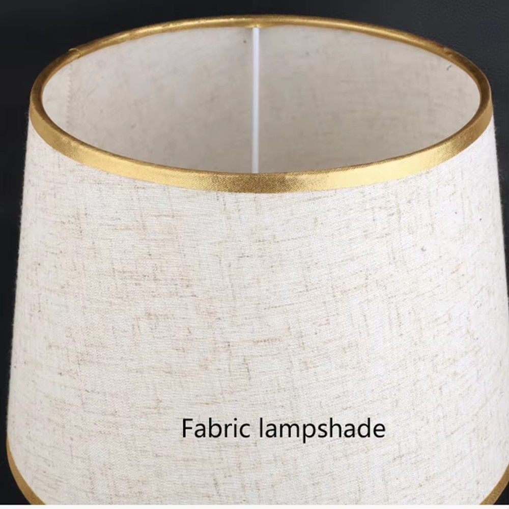 Modern light luxury fabric table lamp, with an off-white fabric lampshade (with gold trim on the top and bottom edges) paired with a black-gold spliced lamp pole and a black disc base, emitting soft and warm light, combining decorativeness and practicality, suitable for lighting and decoration in spaces such as bedroom nightstands, living room side tables, and studies.