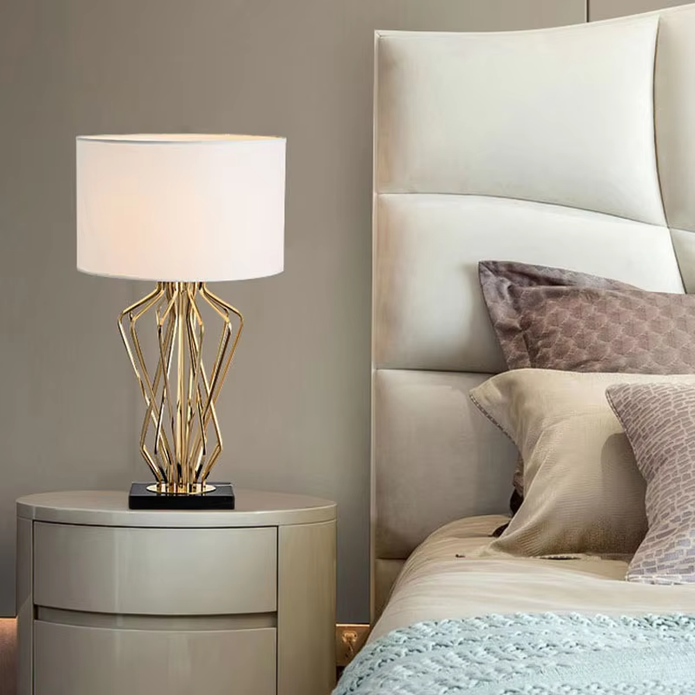 This is a light luxury artistic table lamp, featuring a gold metal geometric hollow lamp body paired with a black marble base and a white fabric lampshade. It emits soft and uniform light, serving both lighting and decorative purposes, suitable for bedroom nightstands, hotel guest rooms, light luxury living room side tables, high-end clubs and other scenarios.
