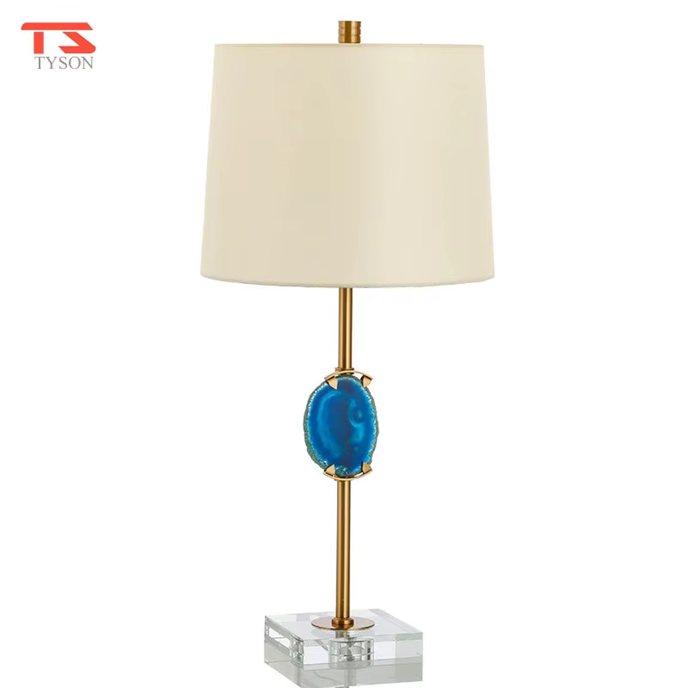 This is a light luxury agate decorative table lamp, with an off-white fabric lampshade paired with a gold metal lamp pole, inlaid with a natural blue agate slice in the middle, and a transparent crystal square base, featuring both decorative and lighting functions, ideal for high-end indoor scenarios such as bedroom nightstands and living room side tables.