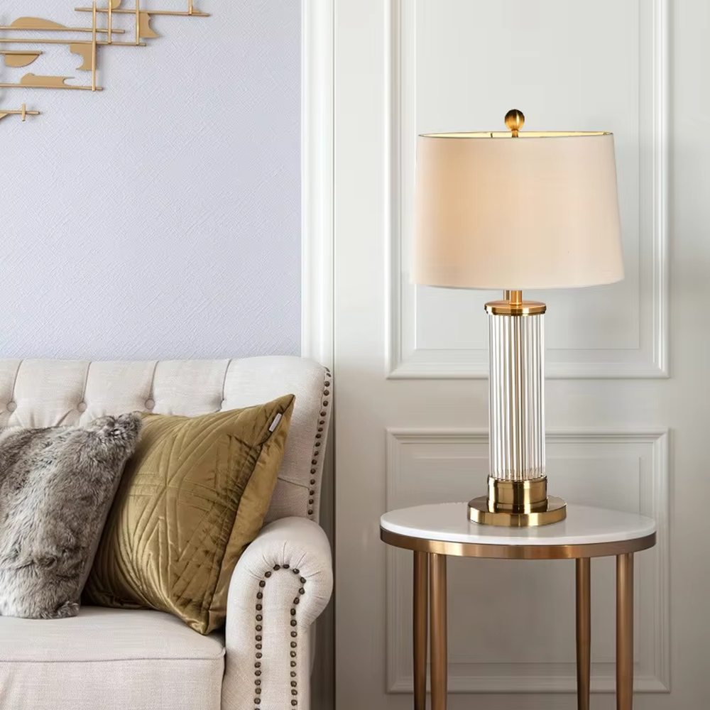 This is a light luxury crystal metal table lamp, with an off-white fabric lampshade edged with gold trim, a lamp post made of vertical striped transparent crystal paired with gold metal splicing, and a gold metal base, featuring both decorative and lighting functions, ideal for high-end indoor scenarios such as bedroom nightstands and living room side tables.