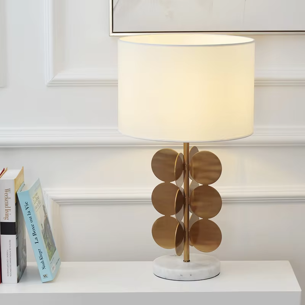This light luxury table lamp features a white fabric cylindrical lampshade on top, a lamp post with a staggered stack of gold metal discs, and a white marble base, serving both lighting and decorative purposes, ideal for living room side tables, bedroom bedside tables, light luxury homestays and other spaces.