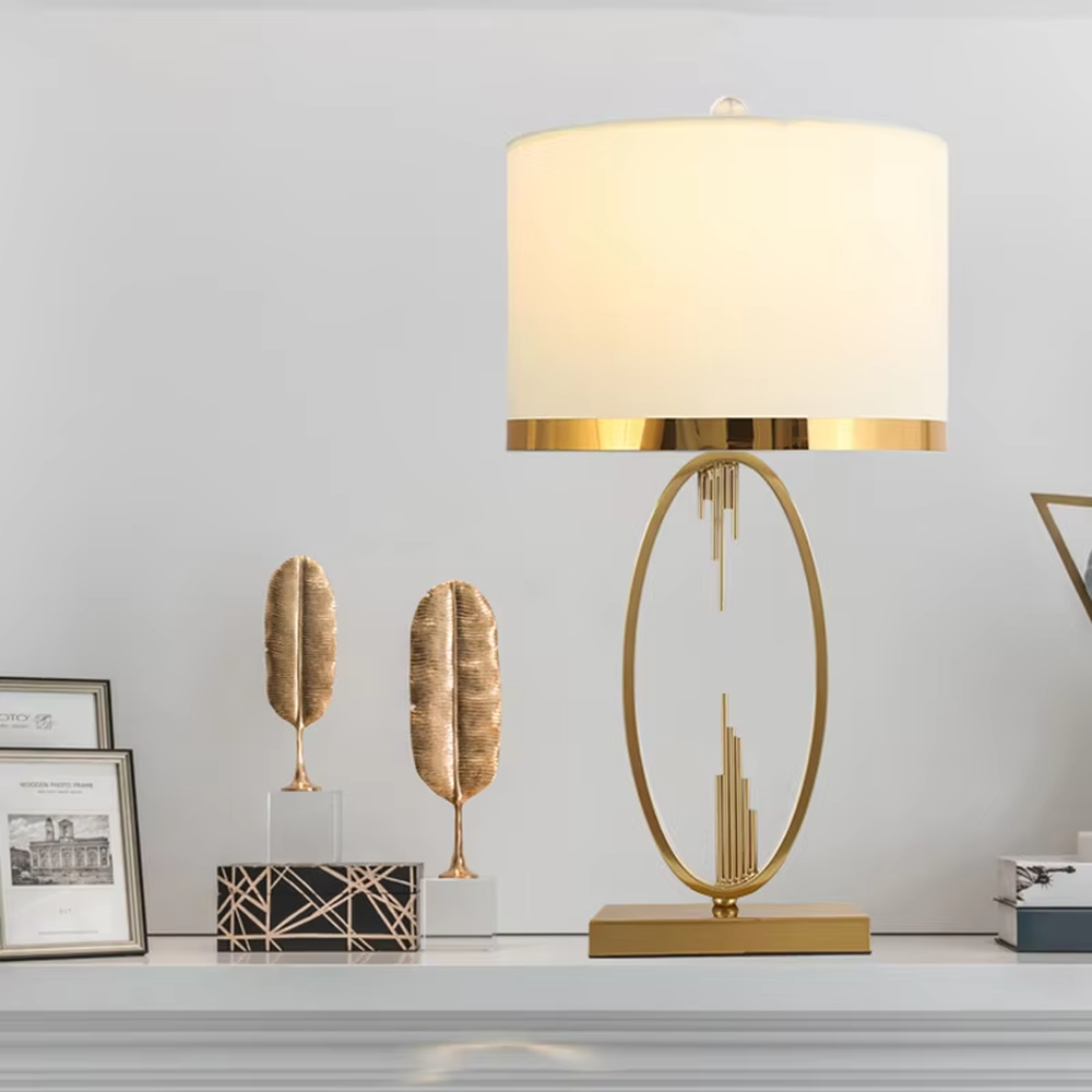 Light luxury style gold metal table lamp, featuring an oval hollow frame with pendant line decorations, paired with an off-white fabric lampshade with a gold trim at the bottom. It emits soft and elegant light, combining artistic decorativeness and lighting practicality, suitable for atmosphere creation and embellishment in bedrooms, living rooms, hotels and other spaces.