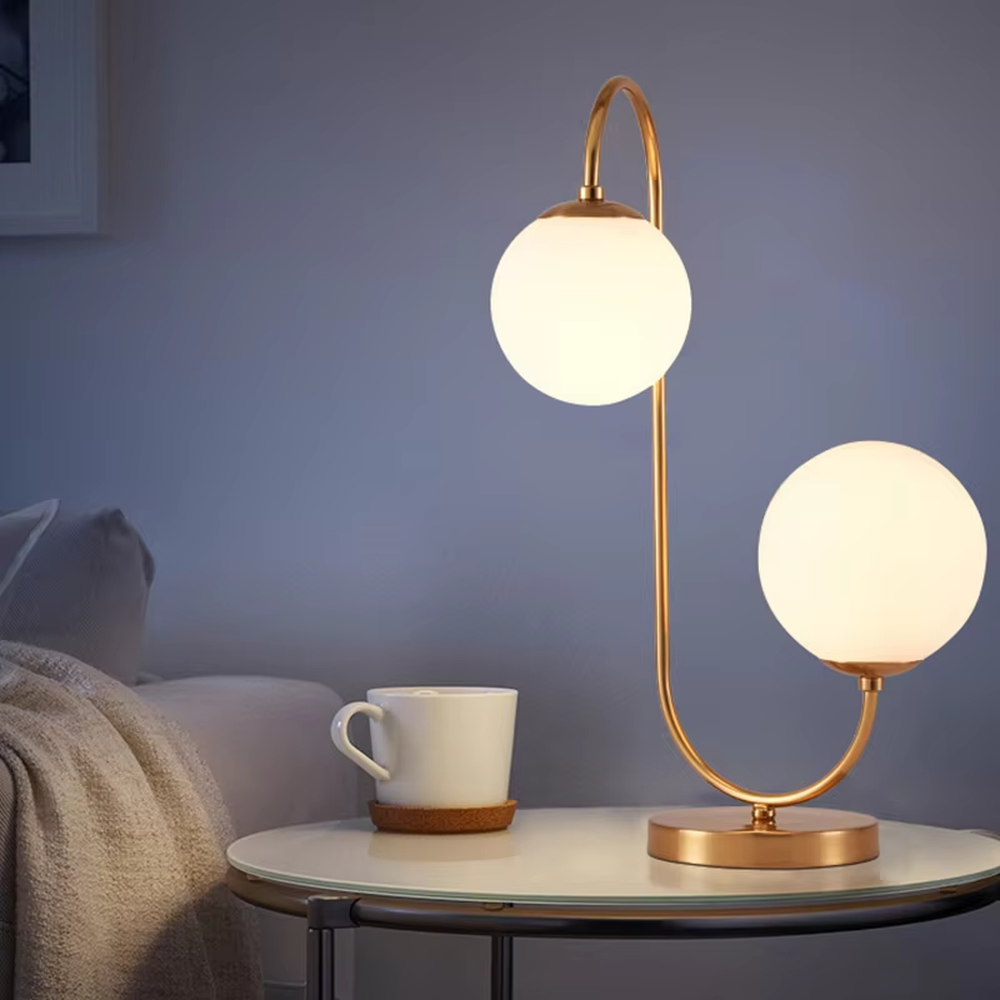 Light luxury double-ball curved table lamp (gold metal curved bracket + double milk-white glass ball lampshades + round metal base), warm light effect, creating a modern and elegant home atmosphere.