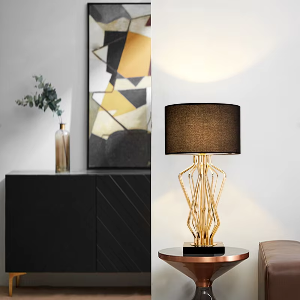 This is a light luxury artistic table lamp, featuring a gold metal geometric hollow lamp body paired with a black marble base and a white fabric lampshade. It emits soft and uniform light, serving both lighting and decorative purposes, suitable for bedroom nightstands, hotel guest rooms, light luxury living room side tables, high-end clubs and other scenarios.