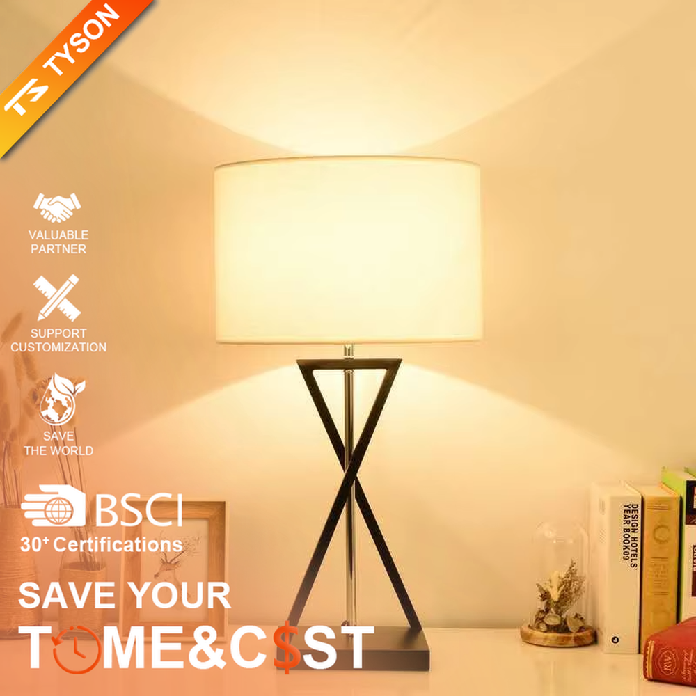 This is a modern minimalist fabric table lamp, featuring an off-white linen lampshade paired with a geometric cross-shaped white and silver dual-color lamp base, which has both lighting and decorative functions, suitable for bedroom nightstands, study desks, hotel guest rooms and other spaces.