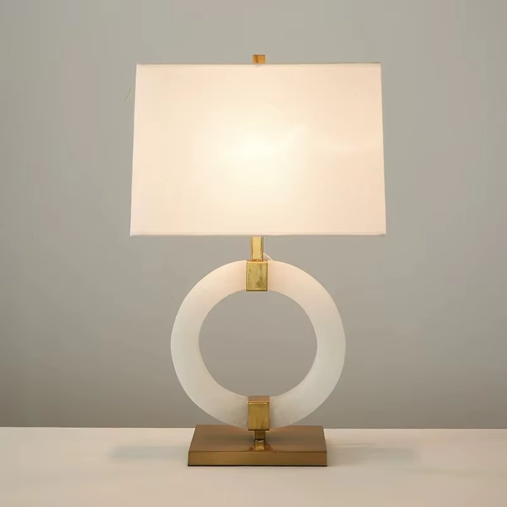 Modern light luxury style table lamp with a white natural stone/acrylic ring paired with gold metal brackets and base, and a rectangular white fabric lampshade. It emits soft and even light, combining decorativeness and practicality, suitable for lighting and decoration in spaces such as bedroom nightstands, living room side tables, and hotel guest rooms.