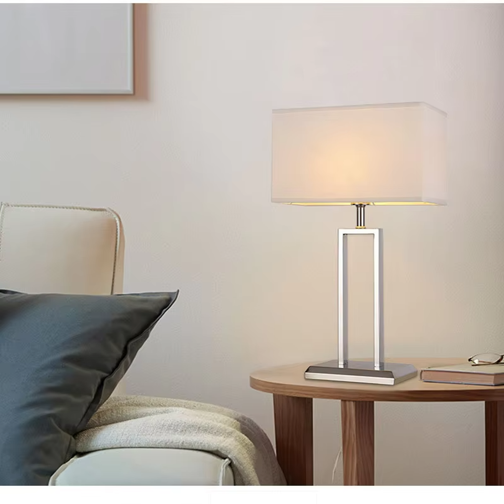 Modern minimalist table lamp with a chrome-plated metal frame and a rectangular off-white fabric lampshade, emitting soft and warm light. It is both decorative and practical, suitable for lighting and decoration in living rooms, bedrooms, studies and other spaces.