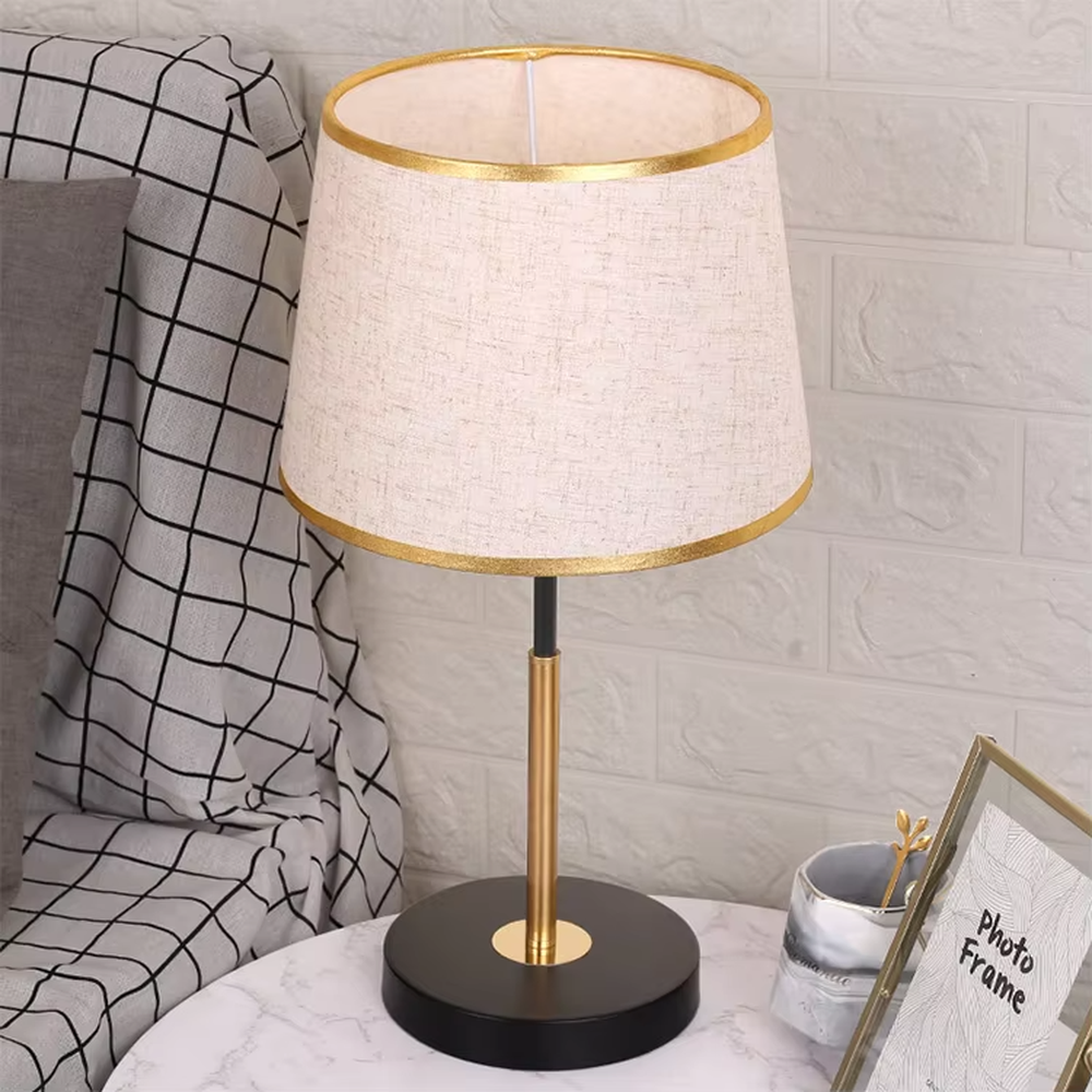 Modern light luxury fabric table lamp, with an off-white fabric lampshade (with gold trim on the top and bottom edges) paired with a black-gold spliced lamp pole and a black disc base, emitting soft and warm light, combining decorativeness and practicality, suitable for lighting and decoration in spaces such as bedroom nightstands, living room side tables, and studies.