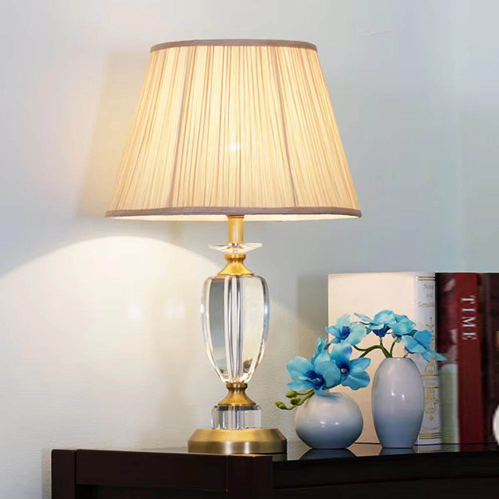 Light luxury crystal table lamp, with a transparent cut crystal column base, gold metal details and a beige pleated fabric lampshade for soft and eye-protective lighting, designed to enhance the elegant atmosphere of spaces, suitable for bedroom nightstands, living room side tables, hotel guest rooms, etc.
