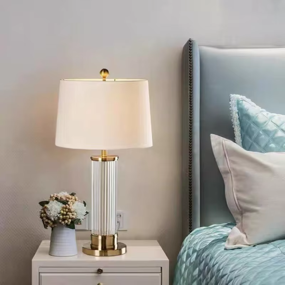 This is a light luxury crystal metal table lamp, with an off-white fabric lampshade edged with gold trim, a lamp post made of vertical striped transparent crystal paired with gold metal splicing, and a gold metal base, featuring both decorative and lighting functions, ideal for high-end indoor scenarios such as bedroom nightstands and living room side tables.
