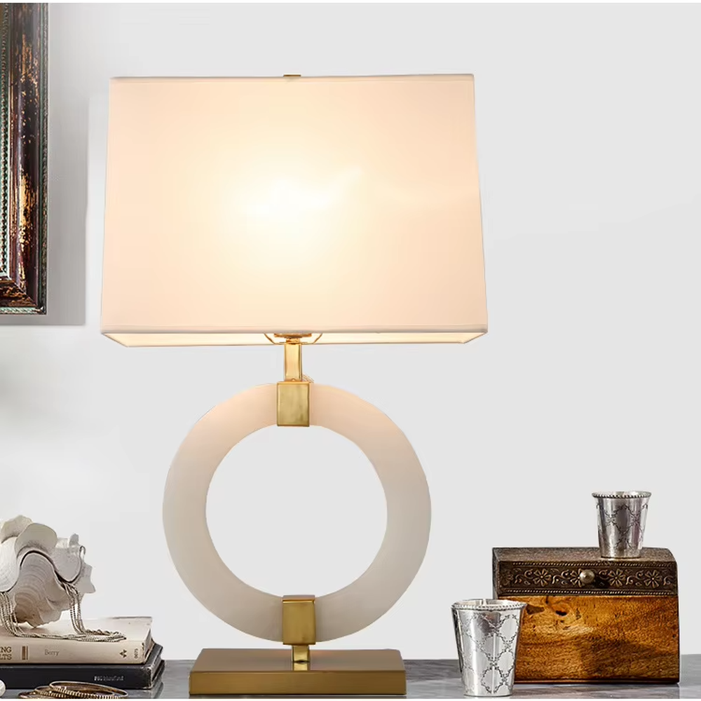 Modern light luxury style table lamp with a white natural stone/acrylic ring paired with gold metal brackets and base, and a rectangular white fabric lampshade. It emits soft and even light, combining decorativeness and practicality, suitable for lighting and decoration in spaces such as bedroom nightstands, living room side tables, and hotel guest rooms.