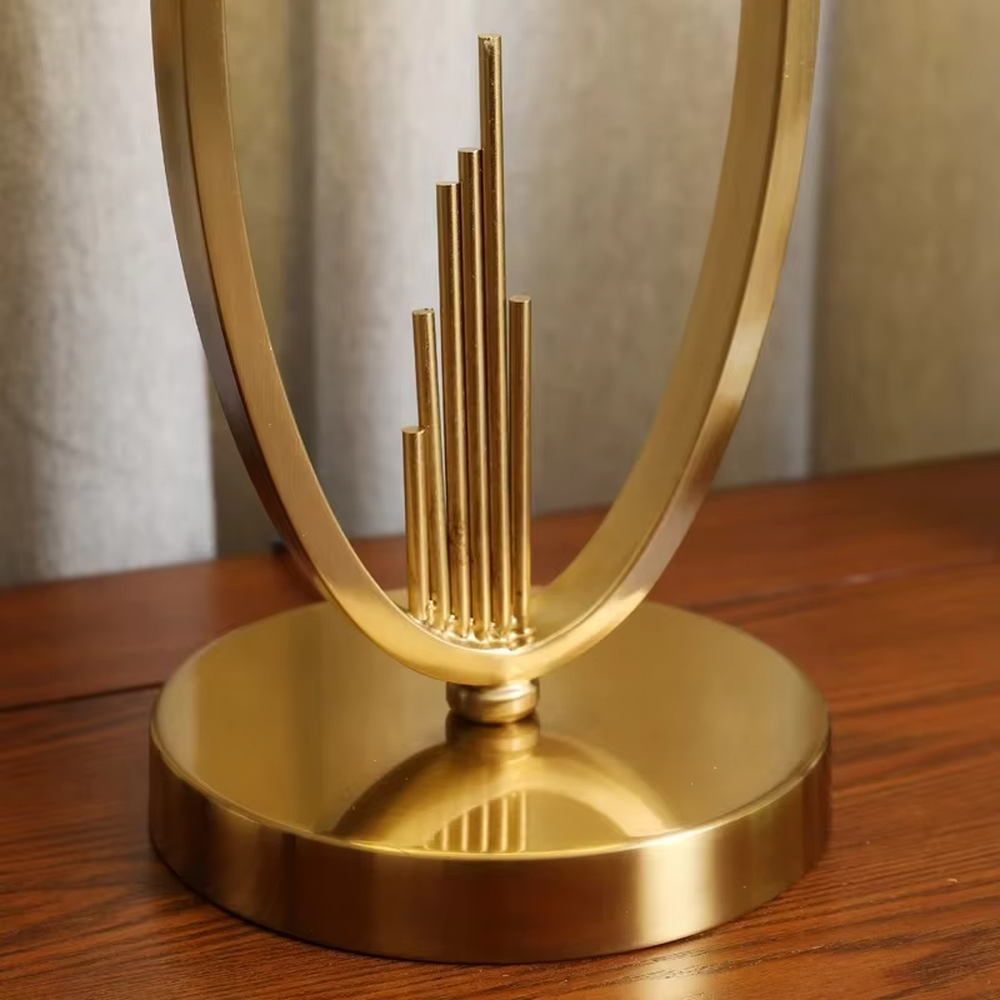 Light luxury style gold metal table lamp, featuring an oval hollow frame with pendant line decorations, paired with an off-white fabric lampshade with a gold trim at the bottom. It emits soft and elegant light, combining artistic decorativeness and lighting practicality, suitable for atmosphere creation and embellishment in bedrooms, living rooms, hotels and other spaces.