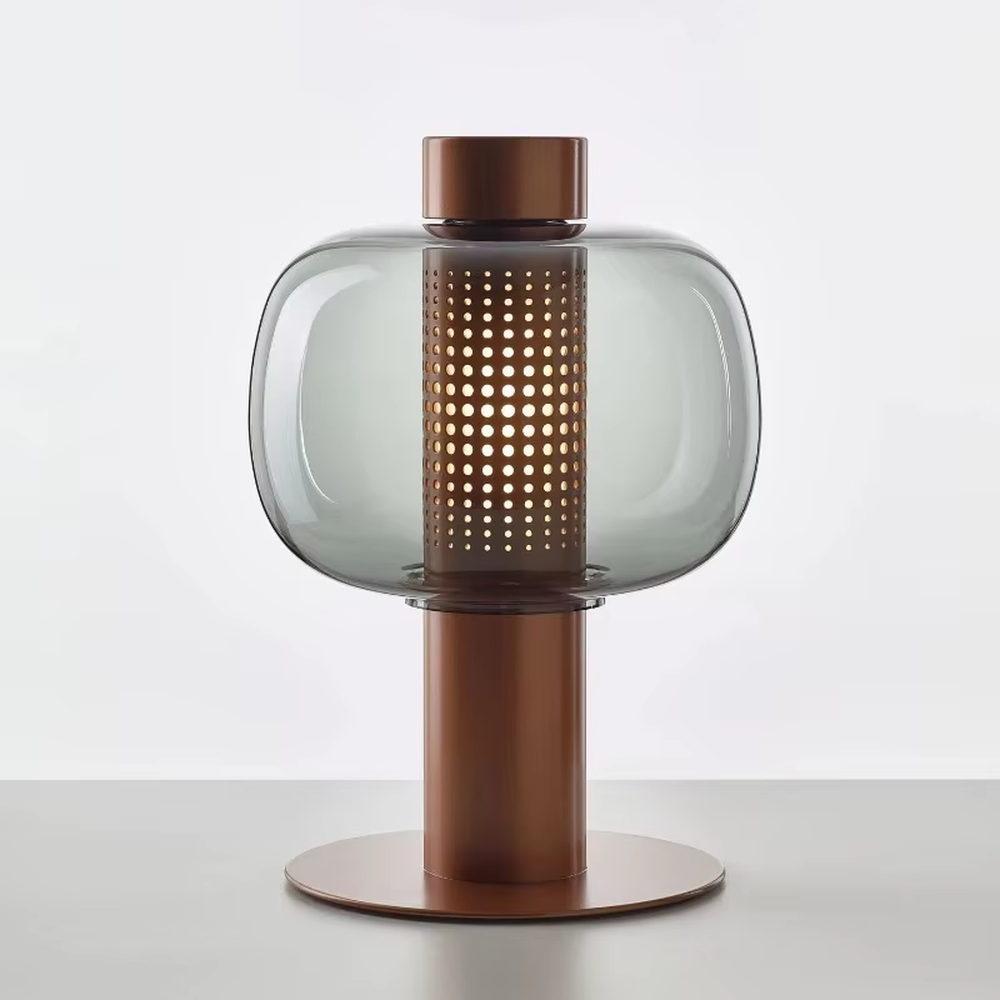 This modern minimalist table lamp features a copper-colored metal frame, paired with an oval smoked glass lampshade and a perforated metal lamp core. Its shape combines roundness with industrial details, and light passes through the perforations and glass to create soft, dappled light and shadow. It serves both lighting and decorative purposes, ideal for bedroom bedside tables, living room side tables, hotel guest rooms, and modern office spaces.