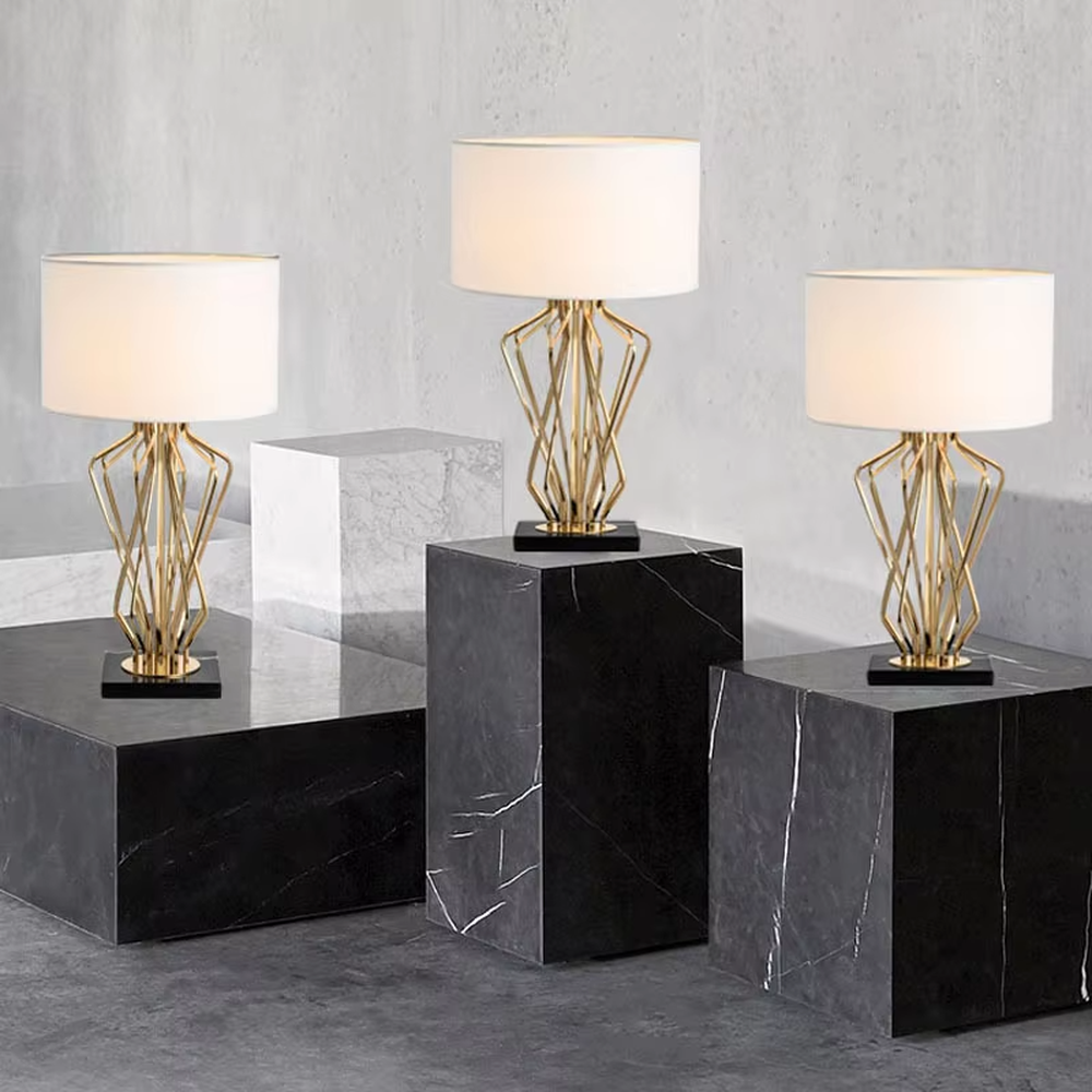This is a light luxury artistic table lamp, featuring a gold metal geometric hollow lamp body paired with a black marble base and a white fabric lampshade. It emits soft and uniform light, serving both lighting and decorative purposes, suitable for bedroom nightstands, hotel guest rooms, light luxury living room side tables, high-end clubs and other scenarios.