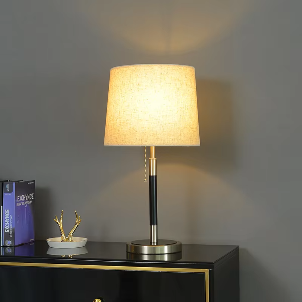This is a minimalist light luxury table lamp, featuring an off-white linen fabric lampshade paired with a black and gold dual-color metal lamp pole, with a pull-chain switch. It emits soft and warm light, serving both lighting and decorative purposes, suitable for bedroom nightstands, living room side tables, studies, hotel guest rooms and other scenarios.