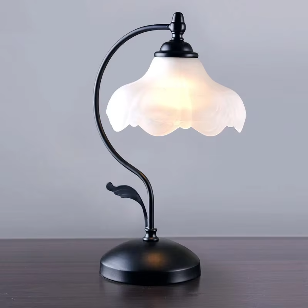 Vintage pastoral style flower-shaped table lamp, with a black wrought iron curved lamp pole with leaf decoration, paired with a white frosted glass petal lampshade. It emits soft and warm light, combining decorativeness and practicality, suitable for lighting and decoration in spaces such as bedroom nightstands, study desks, and B&B guest rooms.