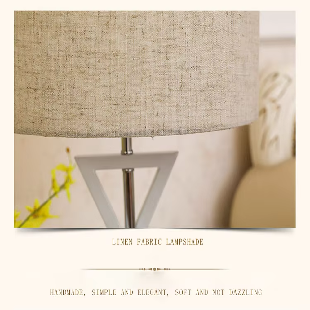 This is a modern minimalist fabric table lamp, featuring an off-white linen lampshade paired with a geometric cross-shaped white and silver dual-color lamp base, which has both lighting and decorative functions, suitable for bedroom nightstands, study desks, hotel guest rooms and other spaces.