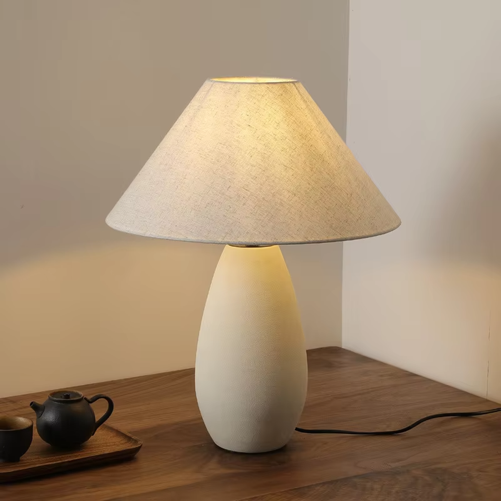 This is a wabi-sabi style minimalist table lamp, featuring an off-white frosted ceramic base paired with a linen fabric lampshade. The light is soft and warm, serving both lighting and decorative purposes, suitable for scenarios pursuing a warm and relaxed feeling such as bedroom nightstands, homestay guest rooms, Japanese-style tea rooms, and wabi-sabi style living rooms.