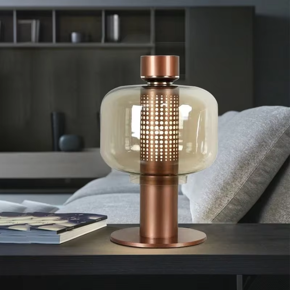 This modern minimalist table lamp features a copper-colored metal frame, paired with an oval smoked glass lampshade and a perforated metal lamp core. Its shape combines roundness with industrial details, and light passes through the perforations and glass to create soft, dappled light and shadow. It serves both lighting and decorative purposes, ideal for bedroom bedside tables, living room side tables, hotel guest rooms, and modern office spaces.