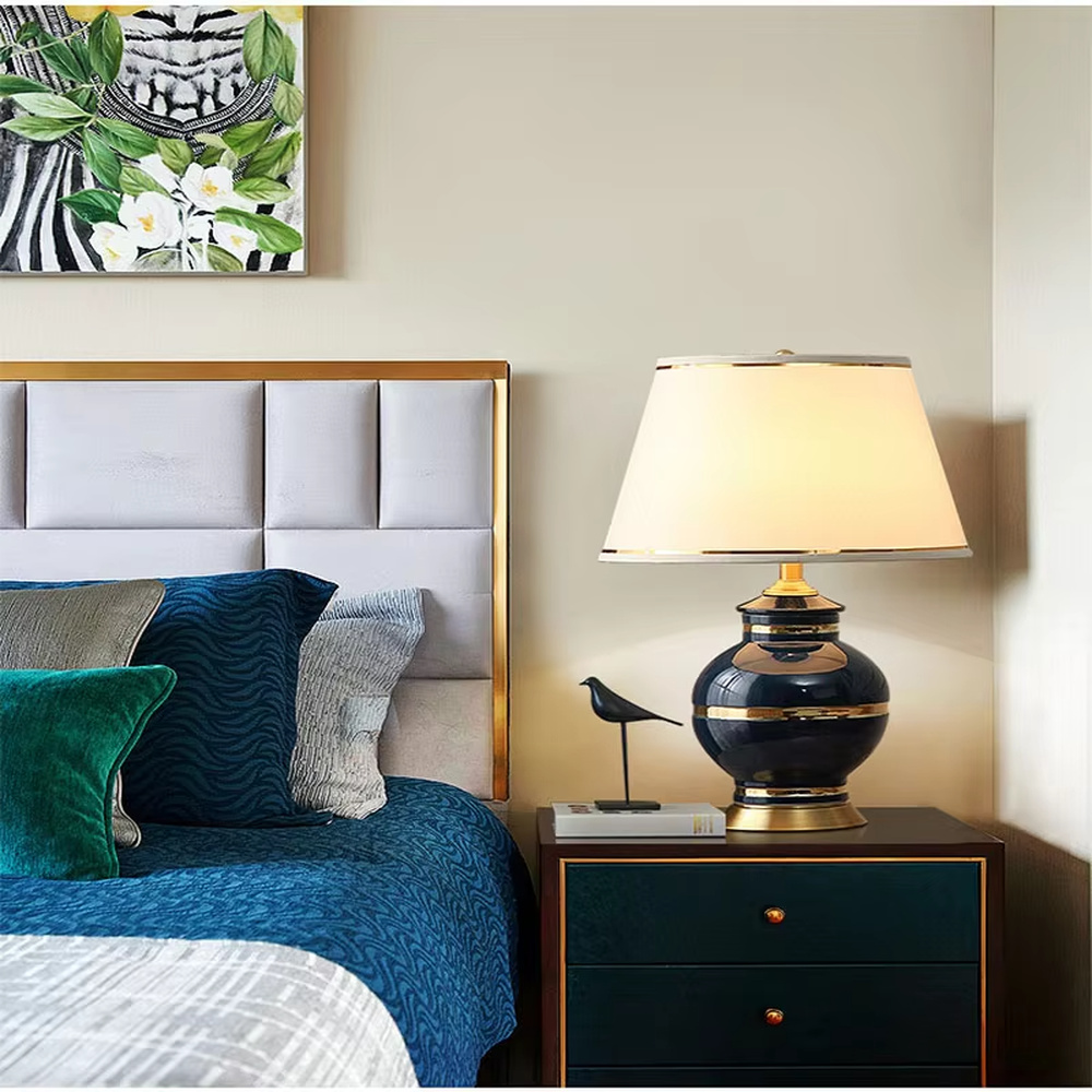 This is a light luxury European-style ceramic table lamp, with a dark blue ceramic base paired with gold metal decorations, and a light blue vertical-striped fabric lampshade edged with a gold ribbon, which has both lighting and decorative functions, suitable for bedroom nightstands, living room side tables, hotel guest rooms and other spaces.
