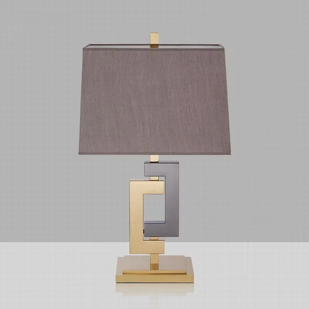 This is a modern light luxury geometric interlocking table lamp, featuring an interlocking geometric lamp body crafted from gold and gray dual-color metal, paired with a dark gray fabric lampshade. It emits soft and uniform light, serving both lighting and decorative purposes, suitable for bedroom nightstands, light luxury living room side tables, hotel guest rooms, showrooms and other scenarios.