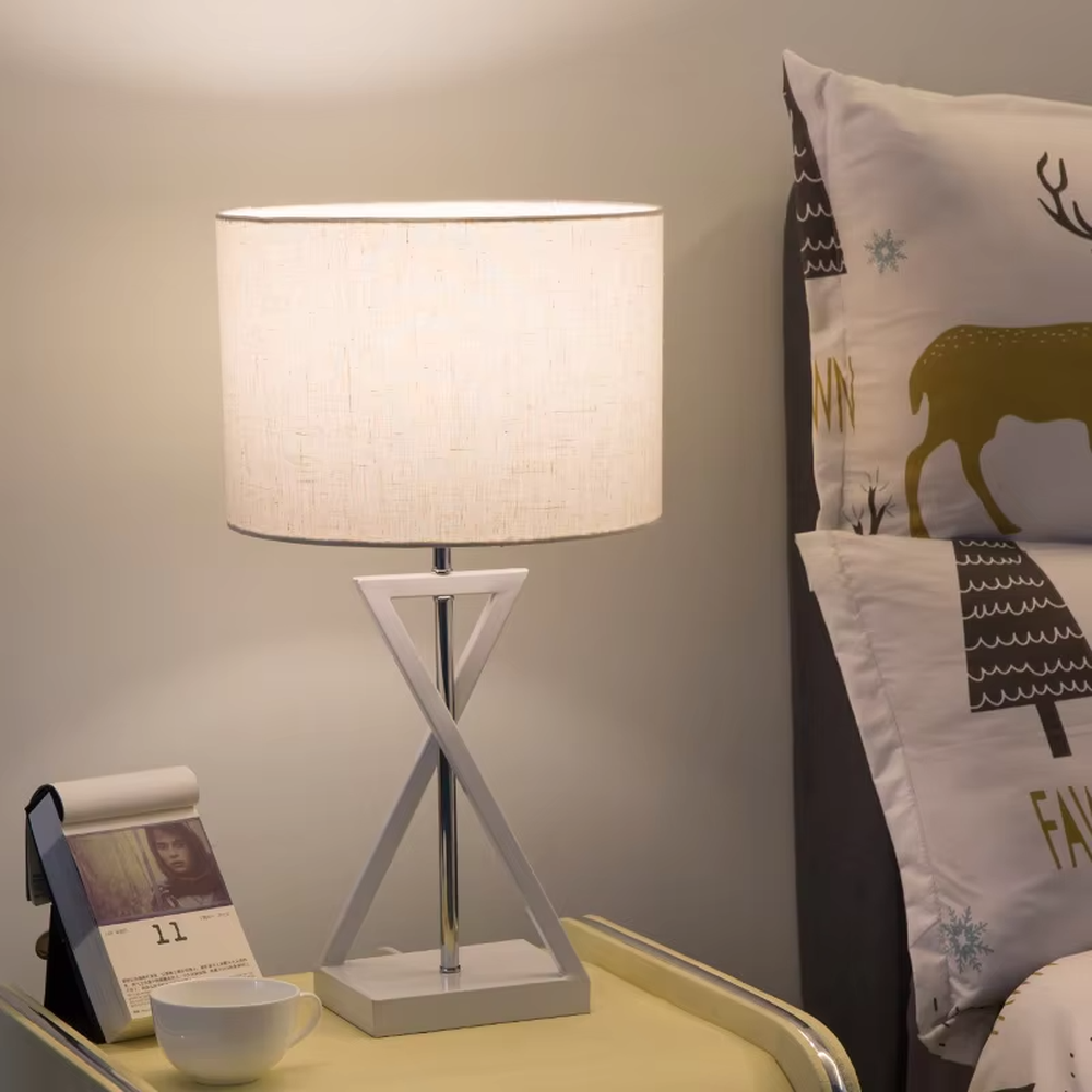 This is a modern minimalist fabric table lamp, featuring an off-white linen lampshade paired with a geometric cross-shaped white and silver dual-color lamp base, which has both lighting and decorative functions, suitable for bedroom nightstands, study desks, hotel guest rooms and other spaces.