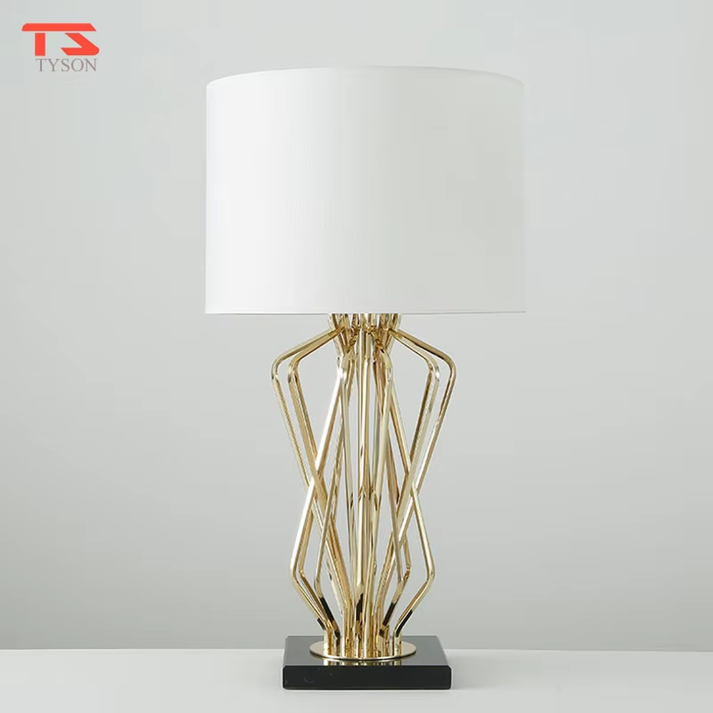 This is a light luxury artistic table lamp, featuring a gold metal geometric hollow lamp body paired with a black marble base and a white fabric lampshade. It emits soft and uniform light, serving both lighting and decorative purposes, suitable for bedroom nightstands, hotel guest rooms, light luxury living room side tables, high-end clubs and other scenarios.