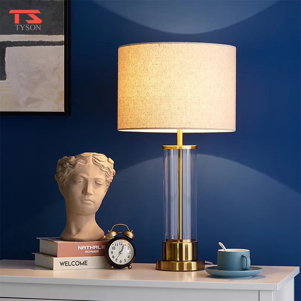 This modern light luxury table lamp features a transparent glass lamp body, paired with gold metal connectors and base, and an off-white fabric cylindrical lampshade that transmits light softly. It serves both lighting and decorative purposes, ideal for light luxury spaces such as bedroom bedside tables, living room side tables, and hotel guest rooms.