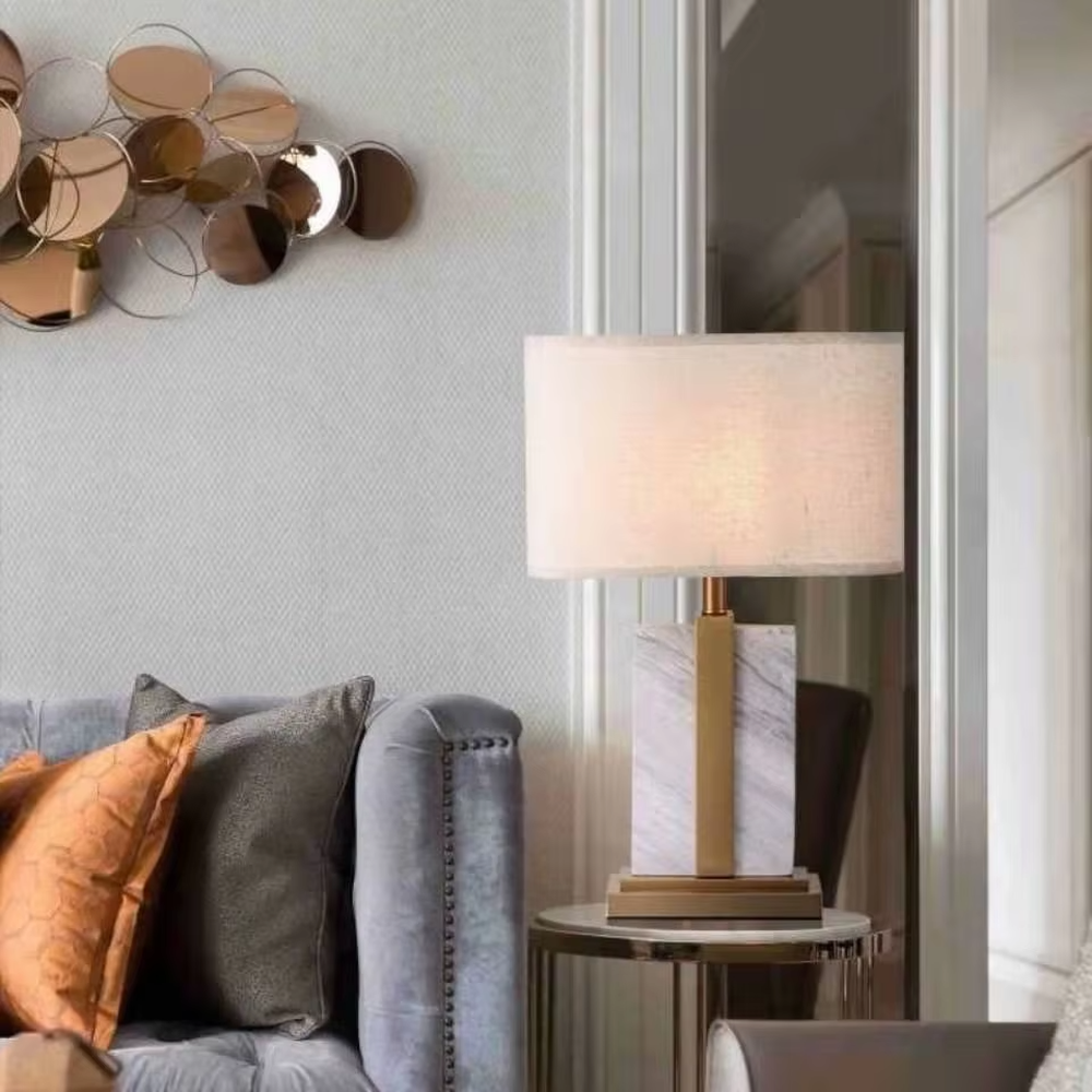 Modern light luxury marble and metal spliced table lamp, with a gray marble lamp body paired with gold metal decorative strips, a light gray fabric rectangular lampshade (with a gold ball decoration on top), emitting soft and elegant light, combining decorativeness and practicality, suitable for lighting and decoration in spaces such as bedroom nightstands, living room side tables, and hotel guest rooms.