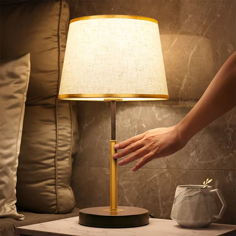 Modern light luxury fabric table lamp, with an off-white fabric lampshade (with gold trim on the top and bottom edges) paired with a black-gold spliced lamp pole and a black disc base, emitting soft and warm light, combining decorativeness and practicality, suitable for lighting and decoration in spaces such as bedroom nightstands, living room side tables, and studies.