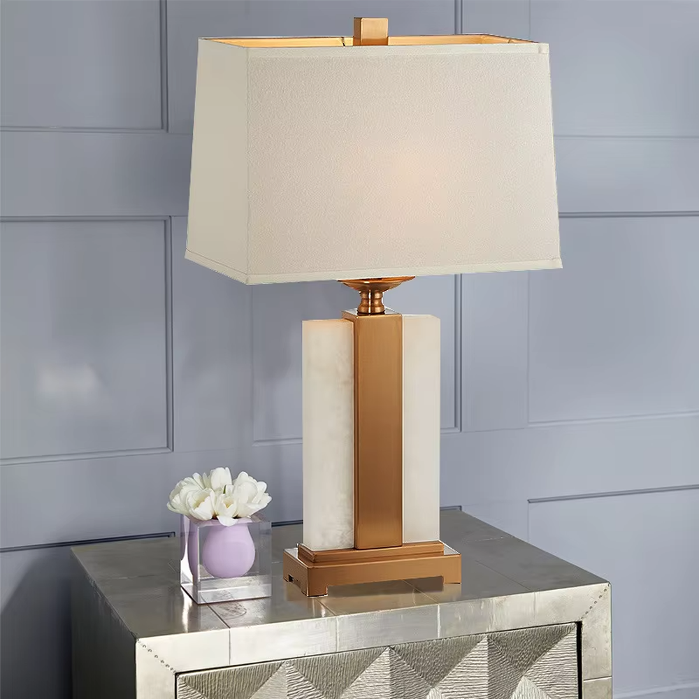 Modern light luxury marble and metal spliced table lamp, with a gray marble lamp body paired with gold metal decorative strips, a light gray fabric rectangular lampshade (with a gold ball decoration on top), emitting soft and elegant light, combining decorativeness and practicality, suitable for lighting and decoration in spaces such as bedroom nightstands, living room side tables, and hotel guest rooms.