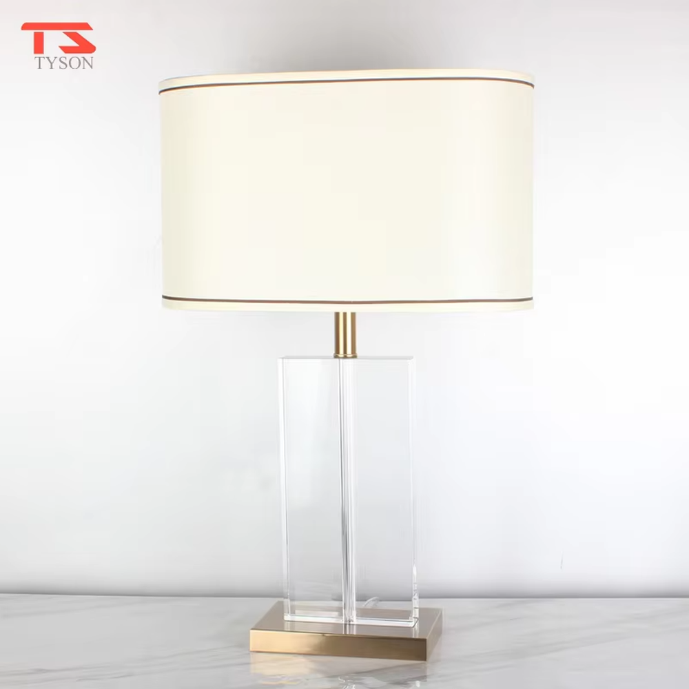 This modern light luxury table lamp features an off-white fabric lampshade (trimmed with a thin gold edge), paired with a transparent crystal square column lamp pole and a gold metal base. The overall style is simple and elegant, with soft eye-protective light, serving both lighting and decorative purposes, suitable for modern light luxury style spaces such as bedrooms, living rooms, and hotel guest rooms.