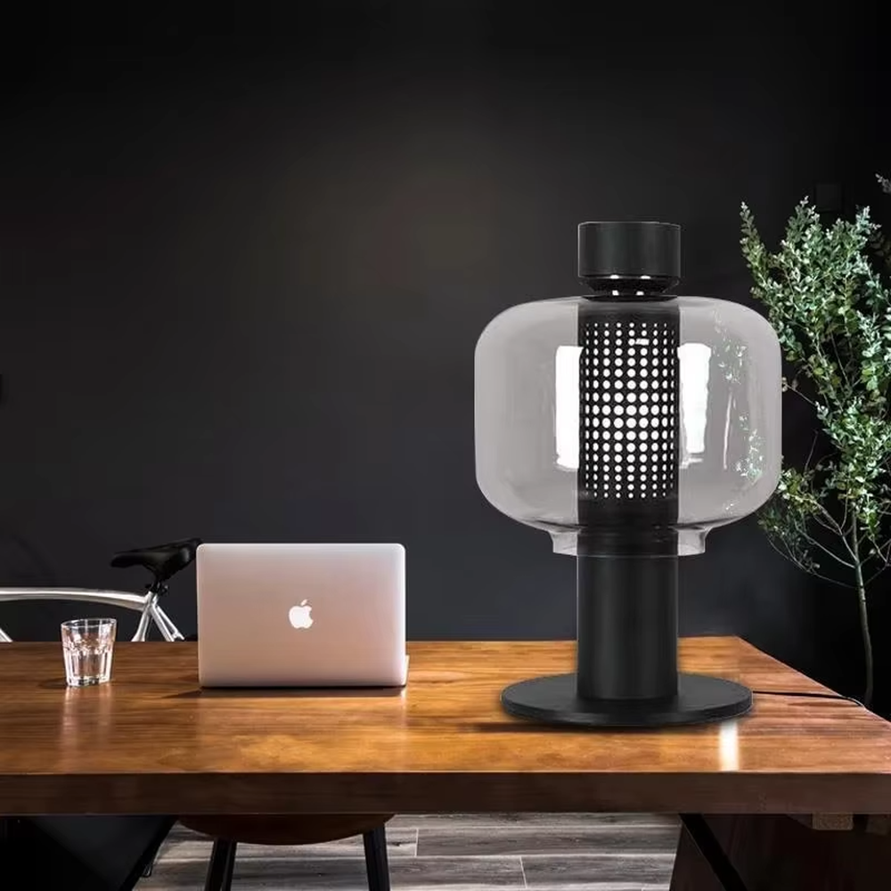 This modern minimalist table lamp features a copper-colored metal frame, paired with an oval smoked glass lampshade and a perforated metal lamp core. Its shape combines roundness with industrial details, and light passes through the perforations and glass to create soft, dappled light and shadow. It serves both lighting and decorative purposes, ideal for bedroom bedside tables, living room side tables, hotel guest rooms, and modern office spaces.