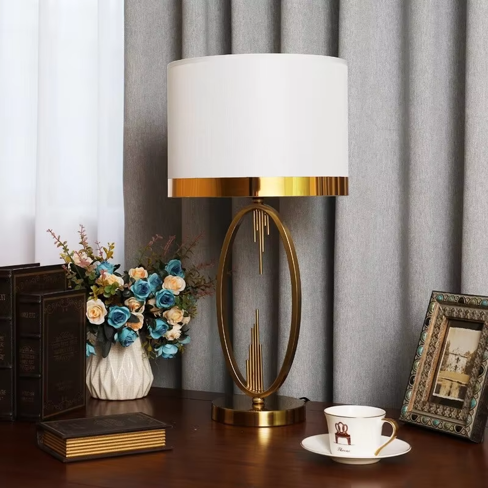 Light luxury style gold metal table lamp, featuring an oval hollow frame with pendant line decorations, paired with an off-white fabric lampshade with a gold trim at the bottom. It emits soft and elegant light, combining artistic decorativeness and lighting practicality, suitable for atmosphere creation and embellishment in bedrooms, living rooms, hotels and other spaces.