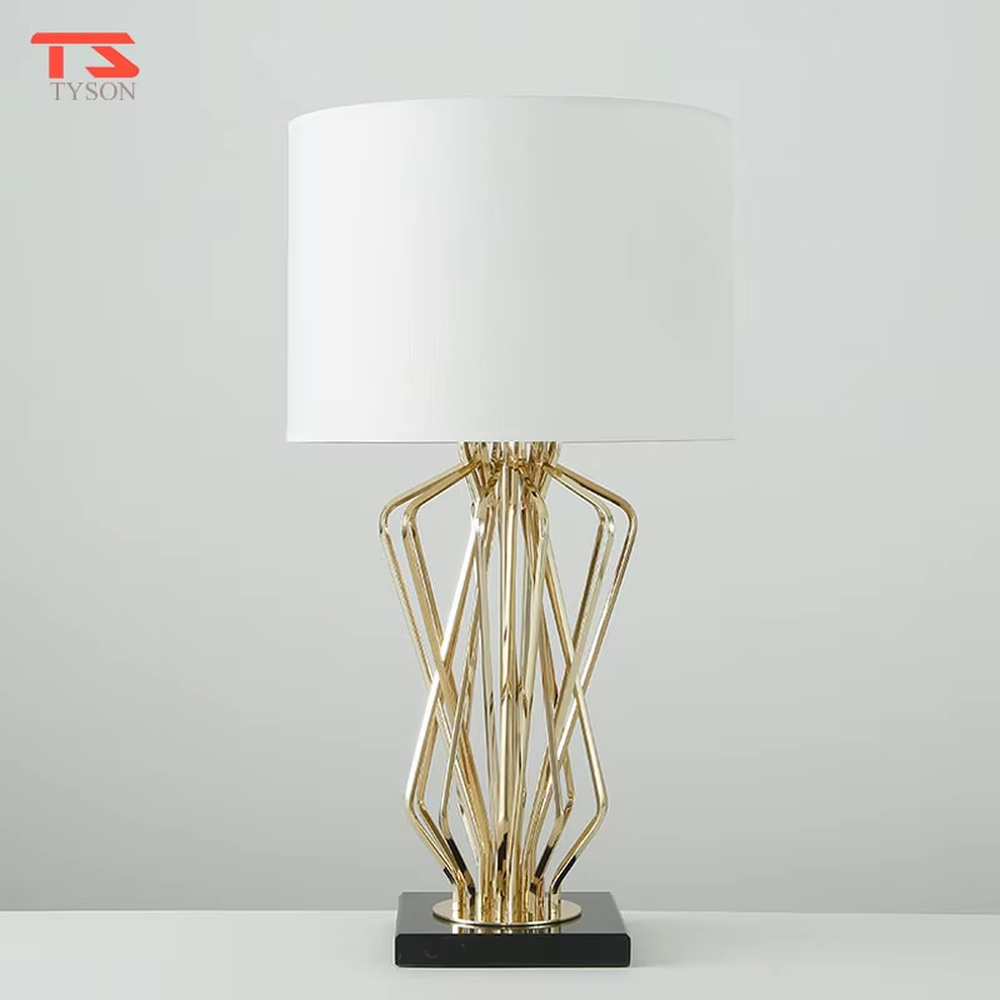 This is a light luxury artistic table lamp, featuring a gold metal geometric hollow lamp body paired with a black marble base and a white fabric lampshade. It emits soft and uniform light, serving both lighting and decorative purposes, suitable for bedroom nightstands, hotel guest rooms, light luxury living room side tables, high-end clubs and other scenarios.