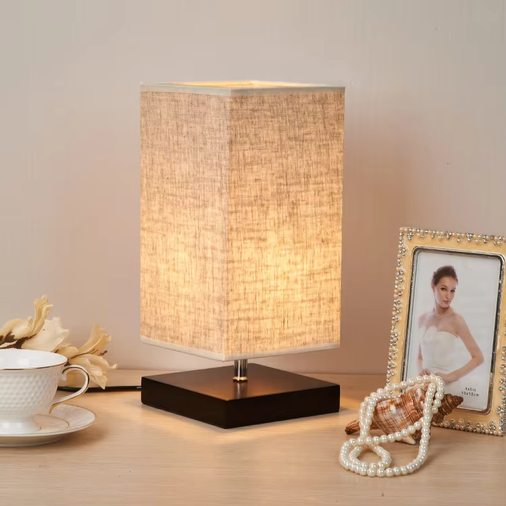This is a minimalist square fabric table lamp, featuring a light beige linen lampshade paired with a black solid wood base. It emits soft and warm light, serving both lighting and decorative purposes, suitable for bedroom nightstands, desks, homestay guest rooms, living room side tables and other scenarios.