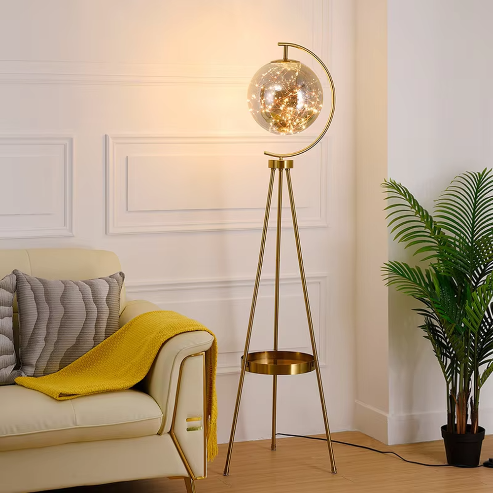 This is a light luxury modern-style golden tripod glass ball floor lamp, with a golden tripod structure, a transparent glass ball (with built-in light string) and a small tray, featuring an exquisite light luxury design, warm lighting. It is used for space lighting and decoration, creating a light luxury and warm atmosphere. Suitable for modern light luxury living rooms, bedroom corners, Nordic-style homes and other places.